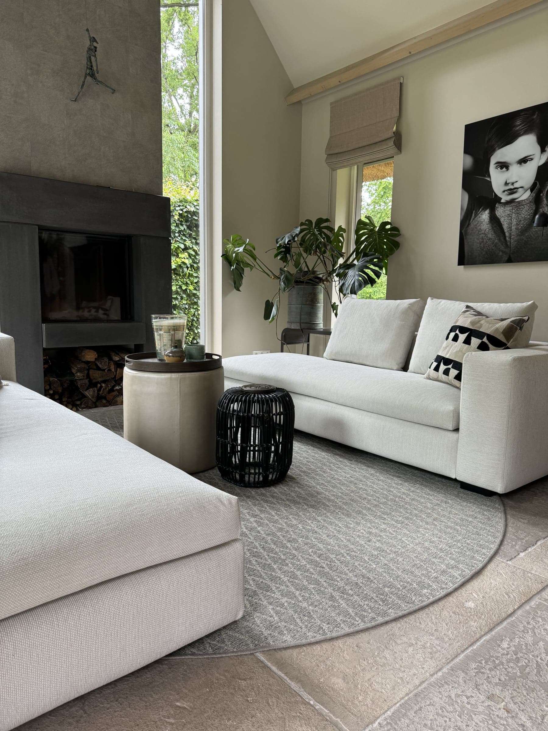 Lichte woonkamer met een witte bank, zwarte gashaard en rond grijs vloerkleed op een natuurstenen vloer in een villa in Waalre