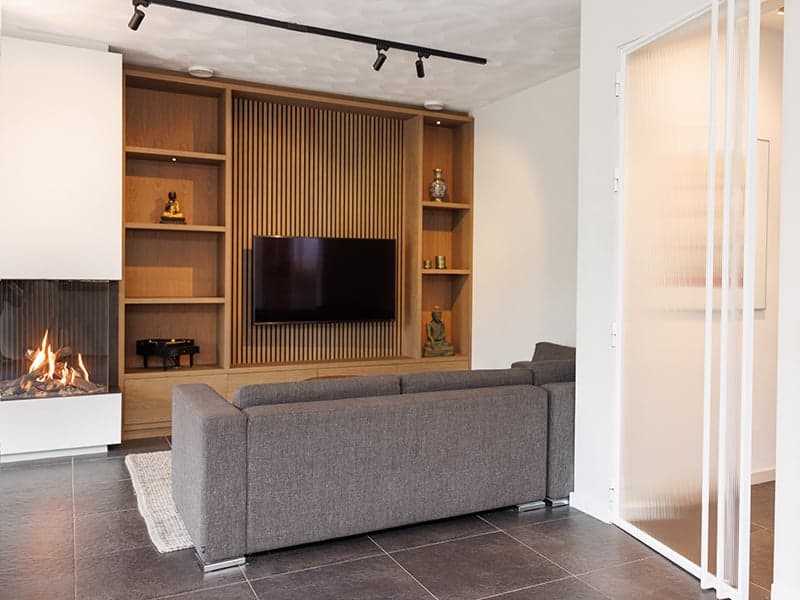 Woonkamer met een grijze bank, houten mediawand met tv en een gashaard op een donkere tegelvloer in een woonhuis in Oisterwijk